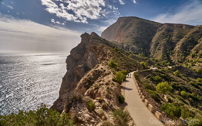 Panorámica del sendero asfaltado que recorre la cresta de los acantilados de la Serra Gelada en la ruta al Faro de l'Albir, con el mar Mediterráneo a la izquierda y la silueta de la montaña al fondo.
