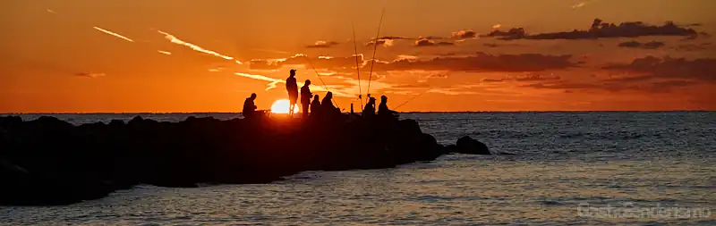 Siluetas de pescadores al amanecer en un espigón de la playa de Port Saplaya, Alboraya, Valencia, con el sol naranja sobre el mar